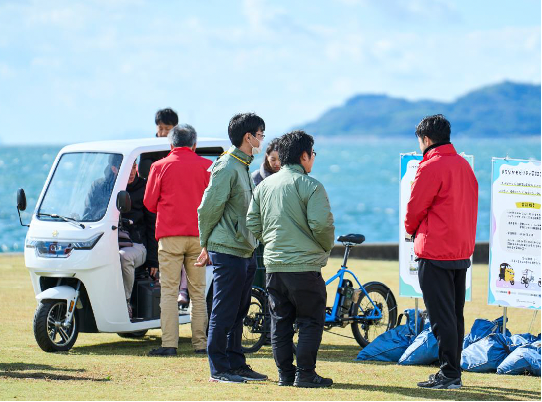 海が見える芝生で「エシカルMaaS」プロジェクトサービスのモビリティが並び説明をしている様子